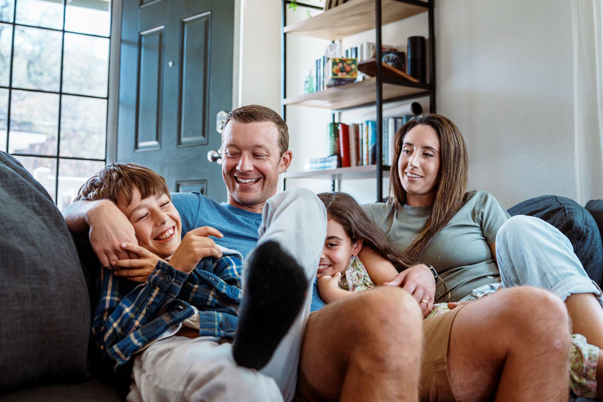 Loving and engaged father and mother sit on the living room sofa with their affectionate elementary age son and daughter as they all cheerfully talk with one another.